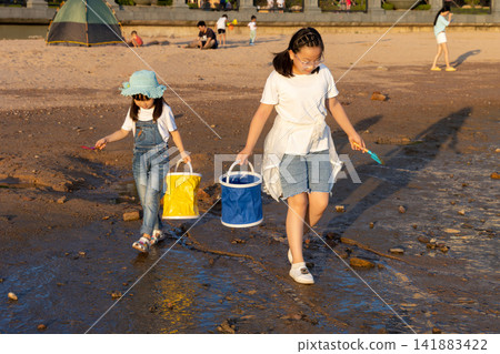 Two young girls carrying colorful water buckets while walking along the wet beach at golden sunset hour. Two young girls carrying colorful water buckets while walking along the wet beach at golden sunset hour. 141883422
