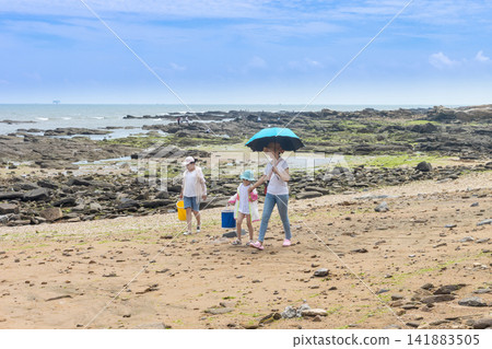 Family walking on a sandy beach during low tide with a blue umbrella for sun protection in summer. Family walking on a sandy beach during low tide with a blue umbrella for sun protection in summer. 141883505