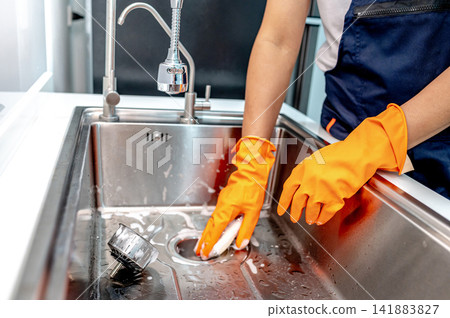 Housekeeper hand wearing orange rubber glove scrubbing on kitchen sink drainer  141883827