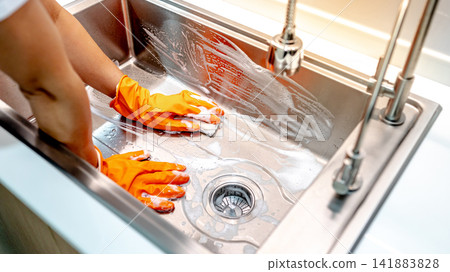 Housekeeper hand wearing orange rubber glove scrubbing on kitchen sink Housekeeper hand wearing orange rubber glove scrubbing on kitchen sink 141883828