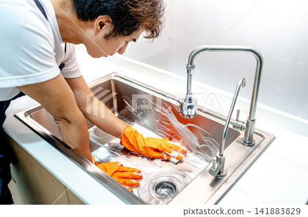 Asian male housekeeper with orange rubber glove scrubbing on kitchen sink Asian male housekeeper with orange rubber glove scrubbing on kitchen sink 141883829