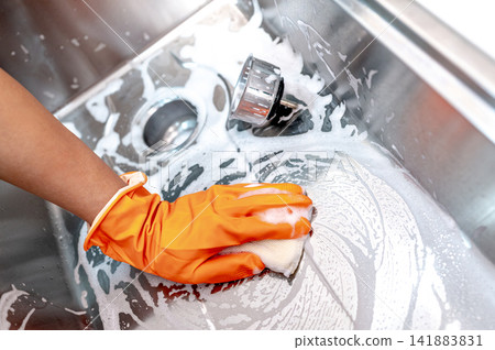 Housekeeper hand wearing orange rubber glove scrubbing on kitchen sink drainer Housekeeper hand wearing orange rubber glove scrubbing on kitchen sink drainer 141883831