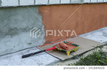 Paint roller rests on tray filled with terracotta paint beside concrete building plinth in outdoor setting Paint roller rests on tray filled with terracotta paint beside concrete building plinth in outdoor setting 141884820