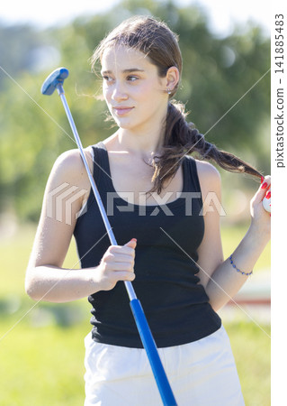 Young girl holds a golf club while playing on a sunny day in the park during the afternoon Young girl holds a golf club while playing on a sunny day in the park during the afternoon 141885483