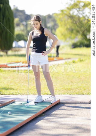 Girl plays mini golf on a sunny day in a park with green grass and trees in the background Girl plays mini golf on a sunny day in a park with green grass and trees in the background 141885484
