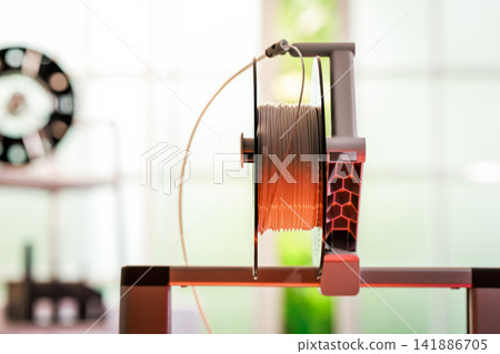 Close-up of a 3d printer filament spool mounted on a printer in a bright workshop, with blurred background and modern technology equipment. Close-up of a 3d printer filament spool mounted on a printer in a bright workshop, with blurred background and modern technology equipment. 141886705