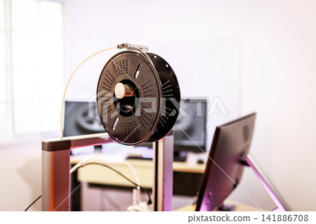 Close-up of a filament spool mounted on a 3d printer in a bright office or workshop, with computer screens in the background Close-up of a filament spool mounted on a 3d printer in a bright office or workshop, with computer screens in the background 141886708