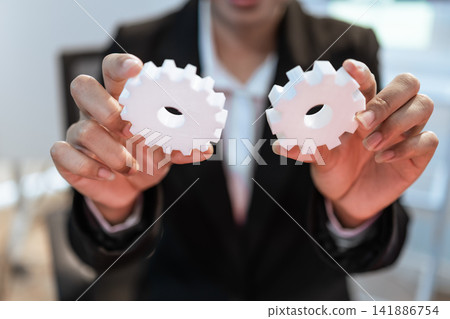 Business person holding two white gear shapes in front of the camera, teamwork, process, planning, connection, and business strategy Business person holding two white gear shapes in front of the camera, teamwork, process, planning, connection, and business strategy 141886754