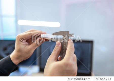 Hands measuring a small white part with a caliper in an office setting, precision, inspection, and quality control Hands measuring a small white part with a caliper in an office setting, precision, inspection, and quality control 141886775