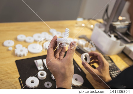 Hands holding 3d printed plastic gears over a workbench with other gear parts and tools for prototype assembly Hands holding 3d printed plastic gears over a workbench with other gear parts and tools for prototype assembly 141886804