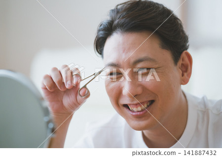 A middle-aged man smiling while trimming his eyebrows with eyebrow scissors - Men's makeup. A middle-aged man smiling while trimming his eyebrows with eyebrow scissors - Men's makeup. 141887432