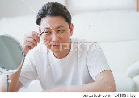 一位笑容可掬的中年男子在桌面鏡子前用眉筆修飾眉毛——男士美容。 一位笑容可掬的中年男子在桌面鏡子前用眉筆修飾眉毛——男士美容。 141887436