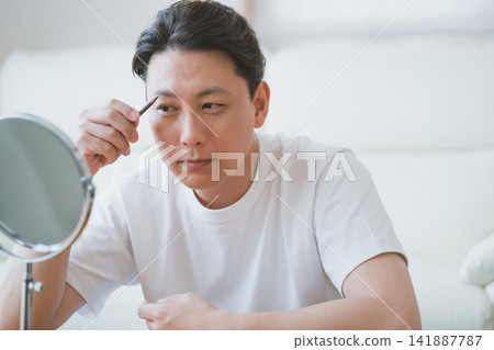 一位中年男子對著桌上鏡用眉筆修飾眉毛——男士美容程序。 一位中年男子對著桌上鏡用眉筆修飾眉毛——男士美容程序。 141887787