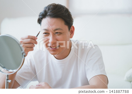 一位中年男子麵帶微笑，在鏡子前用眉筆畫眉——男士化妝。 141887788