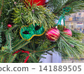 Red Christmas tree ornaments, balls, and a green carnival mask are hanging on the green branches of the fir tree. 141889505