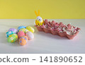 There is a container on the table, and Easter eggs of different colors are lying next to it, including one with bunny ears. 141890652