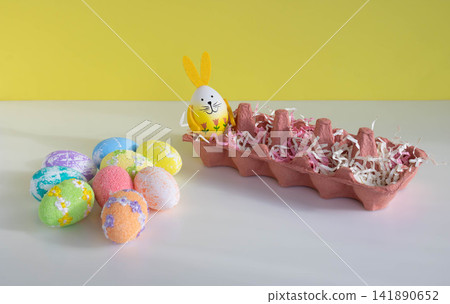 There is a container on the table, and Easter eggs of different colors are lying next to it, including one with bunny ears. 141890652