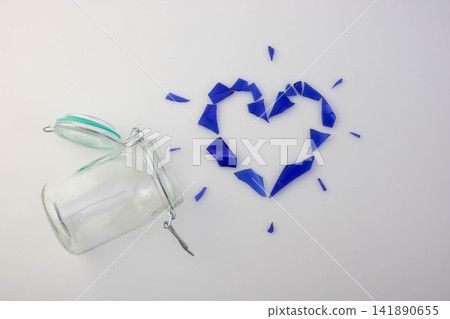 A small glass jar with a lid and a clasp lock fell next to a heart made of blue glass shards on a white background. The concept of love and separation. A small glass jar with a lid and a clasp lock fell next to a heart made of blue glass shards on a white background. The concept of love and separation. 141890655