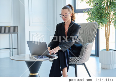 Young focused businesswoman working on laptop at small table in modern office Young focused businesswoman working on laptop at small table in modern office 141891154