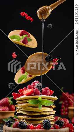 Pancakes fall on a stack of fritters with berries and mint, drizzled with liquid honey from a wooden spoon 141891189