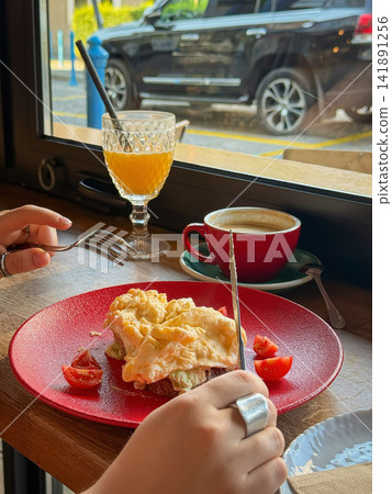 Scrambled Eggs on Toast with cherry tomatoes, coffee and orange drink served on wooden cafe table by window. Casual breakfast scene with hands represents brunch, dining, modern cafe lifestyle Scrambled Eggs on Toast with cherry tomatoes, coffee and orange drink served on wooden cafe table by window. Casual breakfast scene with hands represents brunch, dining, modern cafe lifestyle 141891256