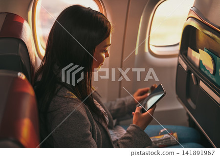 Young Woman Scrolling Smartphone in Profile View Seated by Glowing Airplane Windows in Moody Amber Cabin Light Young Woman Scrolling Smartphone in Profile View Seated by Glowing Airplane Windows in Moody Amber Cabin Light 141891967