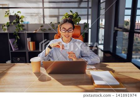 Focused Young Businesswoman Smiling at Laptop Screen at Sunny Modern Office Desk with Coffee Cup Nearby Focused Young Businesswoman Smiling at Laptop Screen at Sunny Modern Office Desk with Coffee Cup Nearby 141891974