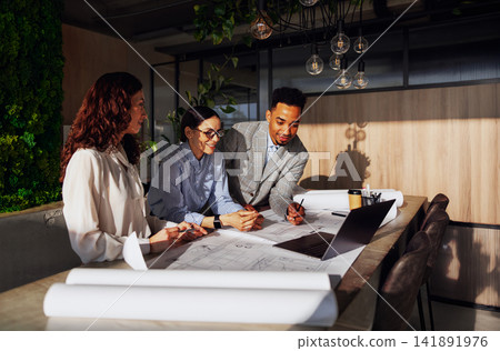Diverse Architecture Team Reviewing Blueprint Plans Together at Biophilic Office Table in Warm Natural Light Diverse Architecture Team Reviewing Blueprint Plans Together at Biophilic Office Table in Warm Natural Light 141891976