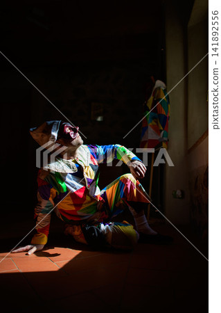 Man in Colorful Harlequin Jester Costume Sitting Indoors in Light Man in Colorful Harlequin Jester Costume Sitting Indoors in Light 141892556