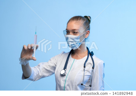 Healthcare professional wearing protective mask, safety goggles, and medical uniform, holding a syringe filled with pink liquid Healthcare professional wearing protective mask, safety goggles, and medical uniform, holding a syringe filled with pink liquid 141892948
