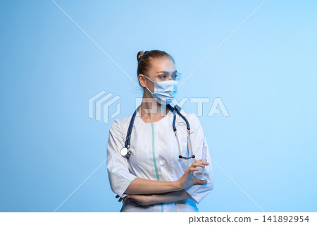 Female medical professional wearing protective mask and glasses, holding a syringe, representing vaccination and healthcare Female medical professional wearing protective mask and glasses, holding a syringe, representing vaccination and healthcare 141892954