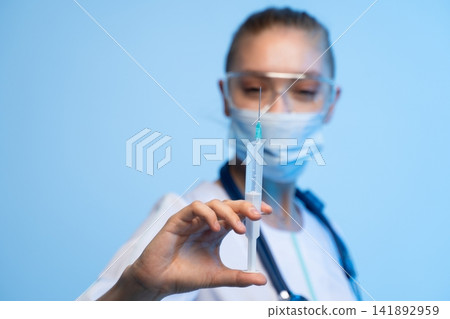Medical professional wearing mask and safety glasses holding a syringe with vaccine for injection or treatment 141892959