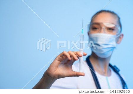 Healthcare professional in mask and safety goggles holding a syringe, preparing injection for vaccination or treatment 141892960