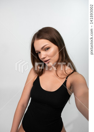 Young woman taking a selfie, posing for a portrait against a white background, wearing a black bodysuit Young woman taking a selfie, posing for a portrait against a white background, wearing a black bodysuit 141893002