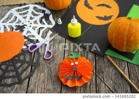 High angle view of a finished handmade paper pumpkin jack-o'-lantern with googly eyes and paper decorations on a wooden table 141893202