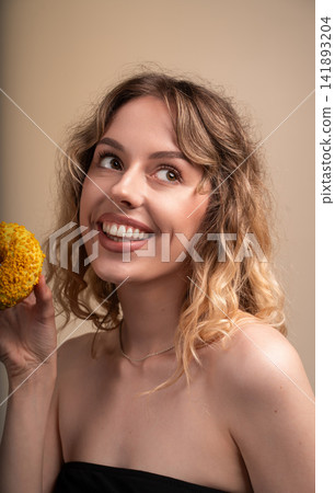 Young woman with curly blonde hair happily holding a yellow doughnut, smiling and looking up, enjoying a sweet snack Young woman with curly blonde hair happily holding a yellow doughnut, smiling and looking up, enjoying a sweet snack 141893204