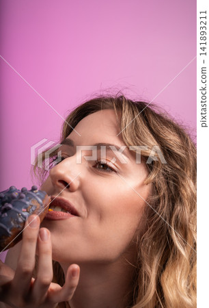 Young woman enjoying delicious sweet donut with purple frosting and sprinkles on vibrant pink background Young woman enjoying delicious sweet donut with purple frosting and sprinkles on vibrant pink background 141893211
