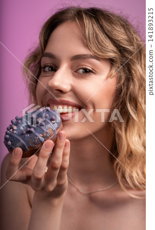 Young woman holding a vibrant purple donut, smiling while taking a bite and enjoying the sweet sugary snack Young woman holding a vibrant purple donut, smiling while taking a bite and enjoying the sweet sugary snack 141893215