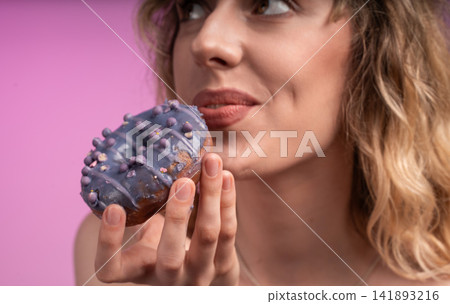 Woman eating sweet purple frosted donut with sprinkles, enjoying dessert and indulging in sugary treat on pink background Woman eating sweet purple frosted donut with sprinkles, enjoying dessert and indulging in sugary treat on pink background 141893216