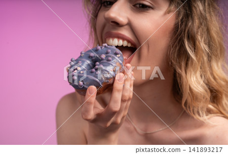 Young woman holding a purple glazed donut, taking a bite while smiling, indulging in a sweet dessert Young woman holding a purple glazed donut, taking a bite while smiling, indulging in a sweet dessert 141893217
