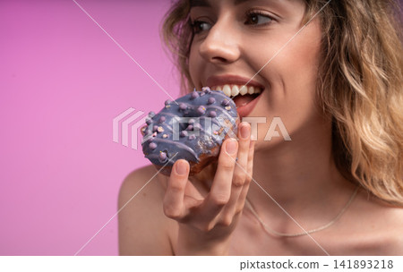 Happy woman enjoying sweet purple glazed donut with sprinkles against a vibrant pink background Happy woman enjoying sweet purple glazed donut with sprinkles against a vibrant pink background 141893218