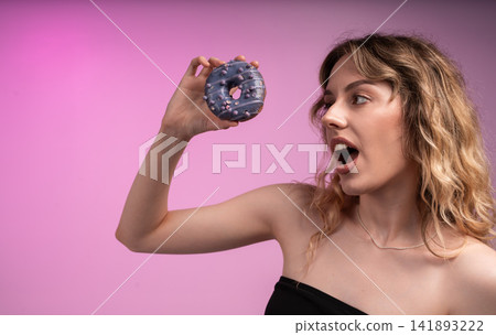 Young woman excitedly anticipating eating purple glazed donut with sprinkles on a vibrant pink background Young woman excitedly anticipating eating purple glazed donut with sprinkles on a vibrant pink background 141893222