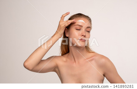 Young woman applying facial moisturizer cream to her forehead with eyes closed for a natural skincare routine experience Young woman applying facial moisturizer cream to her forehead with eyes closed for a natural skincare routine experience 141893281