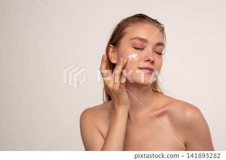 Young woman applying face moisturizer, enjoying a soothing skincare routine for healthy, glowing skin 141893282