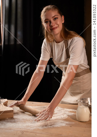 Woman baking homemade dough with rolling pin and flour, preparing bread while smiling in a dark kitchen Woman baking homemade dough with rolling pin and flour, preparing bread while smiling in a dark kitchen 141893322