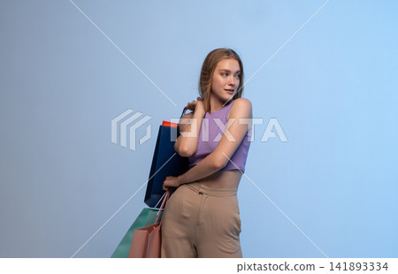 Young woman enjoying shopping, carrying colorful bags, looking away and smiling on a blue background Young woman enjoying shopping, carrying colorful bags, looking away and smiling on a blue background 141893334