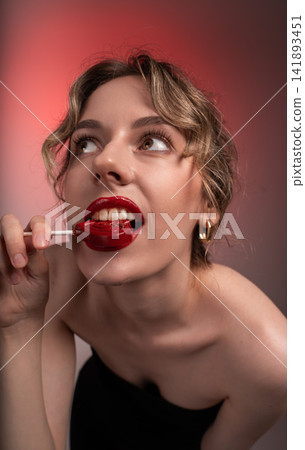 Young woman enjoying sweet red heart lollipop, biting candy with bright red lipstick and expressive eyes looking up Young woman enjoying sweet red heart lollipop, biting candy with bright red lipstick and expressive eyes looking up 141893451