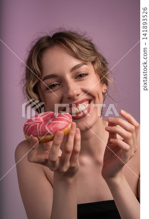 Young woman smiling, holding pink glazed donut, enjoying sweet snack and indulging in dessert Young woman smiling, holding pink glazed donut, enjoying sweet snack and indulging in dessert 141893526