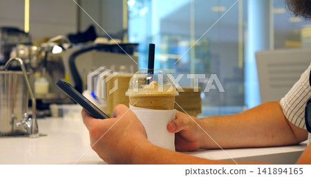 1 Cold drink in clear cup held by person using smartphone at cafe counter with straw and napkin sleeve, soft focus background, casual refreshment and mobile payment context, hand near checkout screen 1 Cold drink in clear cup held by person using smartphone at cafe counter with straw and napkin sleeve, soft focus background, casual refreshment and mobile payment context, hand near checkout screen 141894165