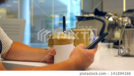 Hands holding cold drink and smartphone at cafe counter, clear cup with dome lid and black straw, iced beverage wrapped in napkin beside blurred espresso machines and bright interior Hands holding cold drink and smartphone at cafe counter, clear cup with dome lid and black straw, iced beverage wrapped in napkin beside blurred espresso machines and bright interior 141894166